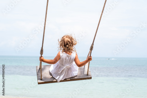 A little girl with curly hair in a white dress is swinging on a swing on a sandy beach by the sea, ocean. Sea holidays, travel and beach holidays with children © Julia