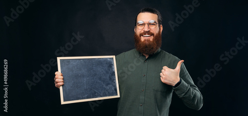 Smiling bearded man is holding a black board while showing thumb up. Panorama studio shot over black background.