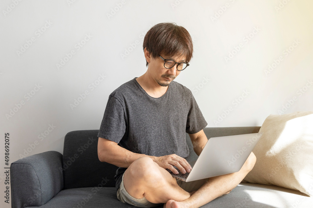 Adult Asian man working with laptop computer and relax on sofa in living room, freelance man using notebook on couch with comfort, new normal, lifestyle concept. Adult Asian man working with laptop computer and relax on sofa in living room, freelance man using notebook on couch with comfort, new normal, lifestyle concept.