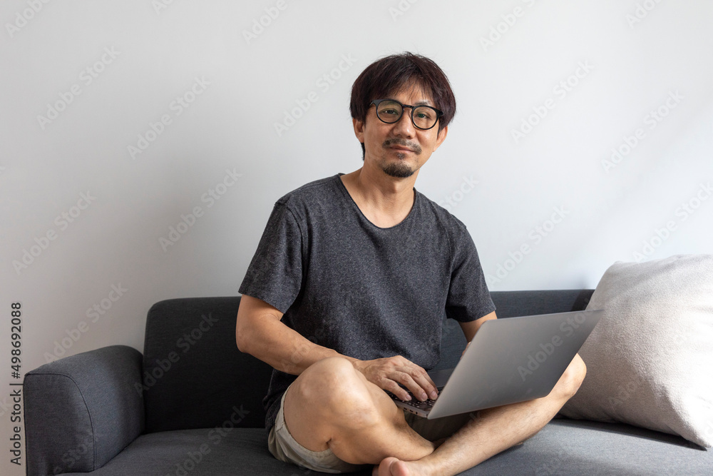 Adult Asian man working with laptop computer and relax on sofa in living room, freelance man using notebook on couch with comfort, new normal, lifestyle concept. Adult Asian man working with laptop computer and relax on sofa in living room, freelance man using notebook on couch with comfort, new normal, lifestyle concept.