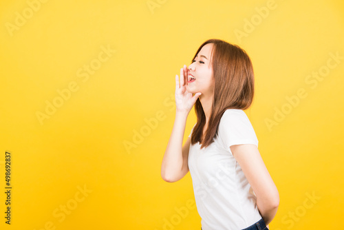 Asian happy portrait beautiful cute young woman teen standing wear t-shirt hand on mouth talking whispering secret rumor looking to side isolated  studio shot on yellow background with copy space