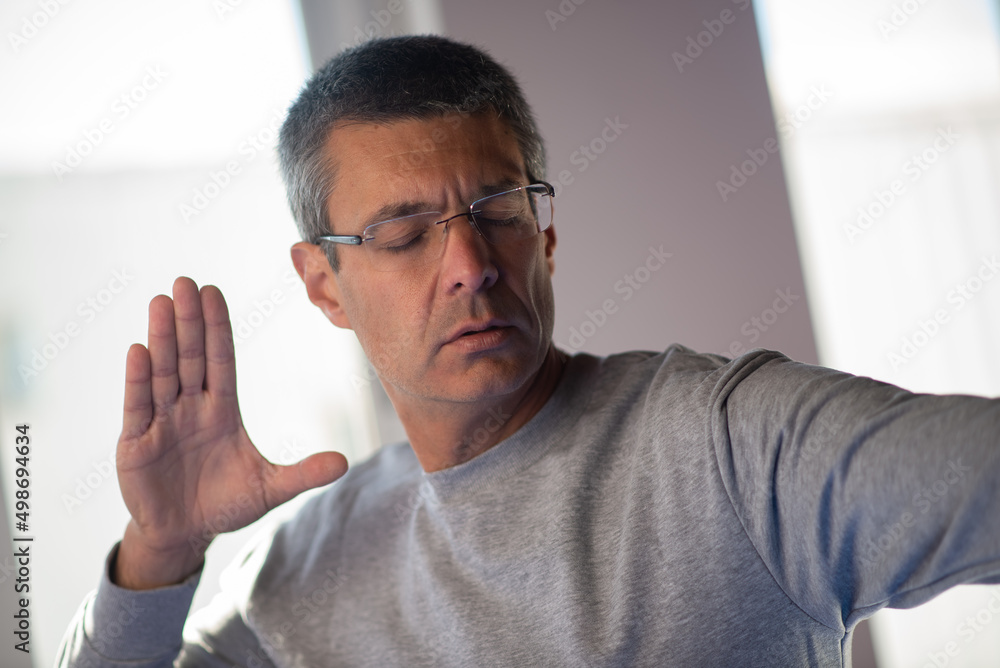 Aged man with closed eyes doing taichi exercise. Concentrated mature man in gray sweater and eyeglasses moving arms in gym. Martial art concept Aged man with closed eyes doing taichi exercise. Concentrated mature man in gray sweater and eyeglasses moving arms in gym. Martial art concept