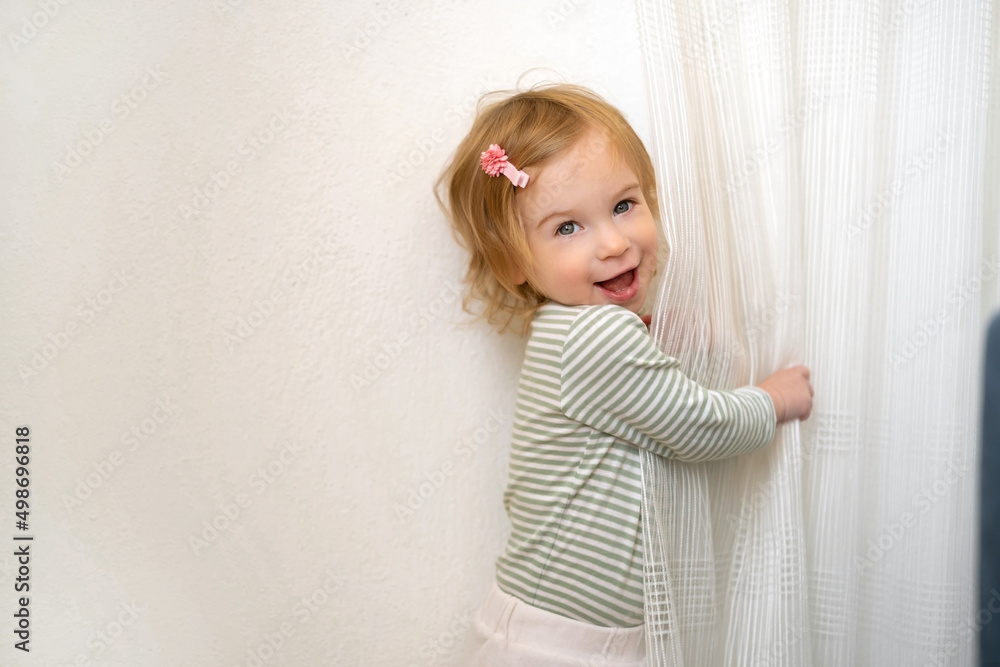 Cute caucasian blonde baby girl of 1,2 year old playing with white curtains,having fun at home in light interior looking at camera.Kid, child,toddler games indoors,laughing,smiling infant.Copy space Cute caucasian blonde baby girl of 1,2 year old playing with white curtains,having fun at home in light interior looking at camera.Kid, child,toddler games indoors,laughing,smiling infant.Copy space