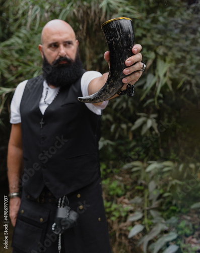 Image of a drinking horn being holding by a bearded man dressed with waistcoat and male skirt who is in a place with plants and streams