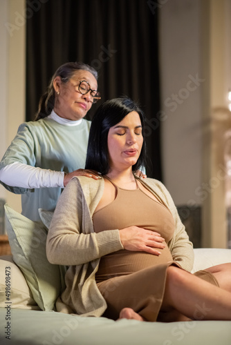 Focused pregnant woman and midwife at home. Woman in casual clothes sitting on bed, Asian doula massaging shoulders. Pregnancy, medicine, home birth concept