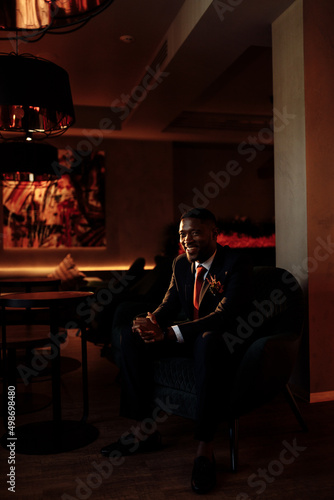 Portrait of a smiling handsome African American business man. Attractive charming man looks at the camera, smiles, sits on the couch. A young guy in a stylish suit looks at the camera with a smile.