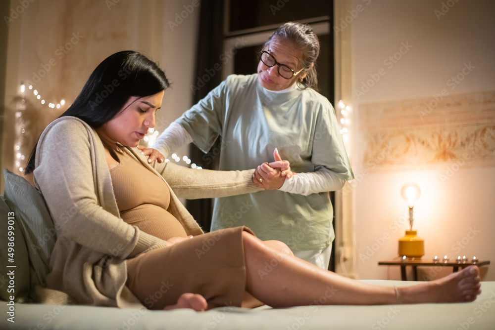 Obraz premium Side view of expecting woman and midwife at home. Woman in casual clothes sitting on bed, Asian doula holding hand. Pregnancy, medicine, home birth concept