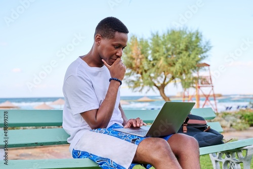 Young serious male freelancer relaxing on beach, sitting on bench using laptop