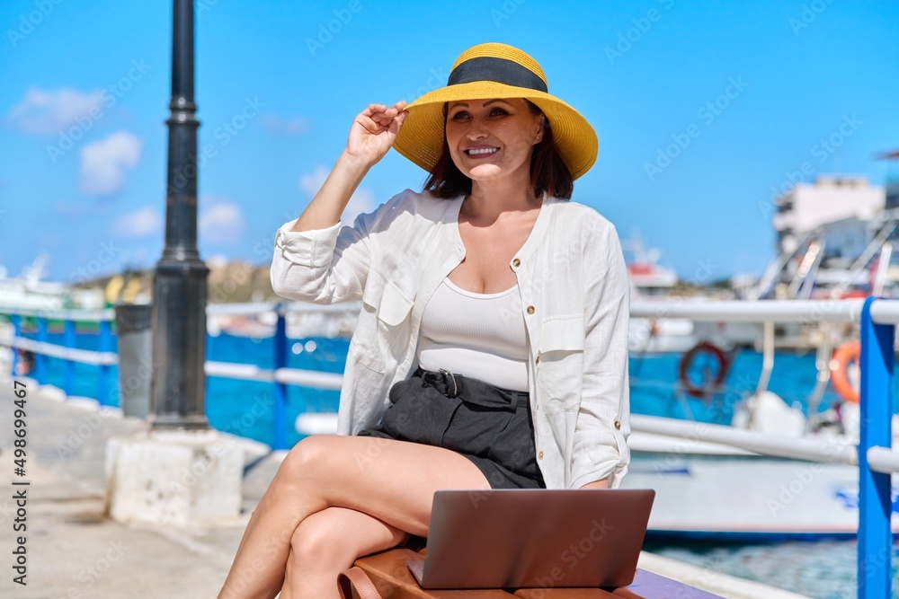 Outdoor, middle aged woman in straw hat sitting on bench using laptop