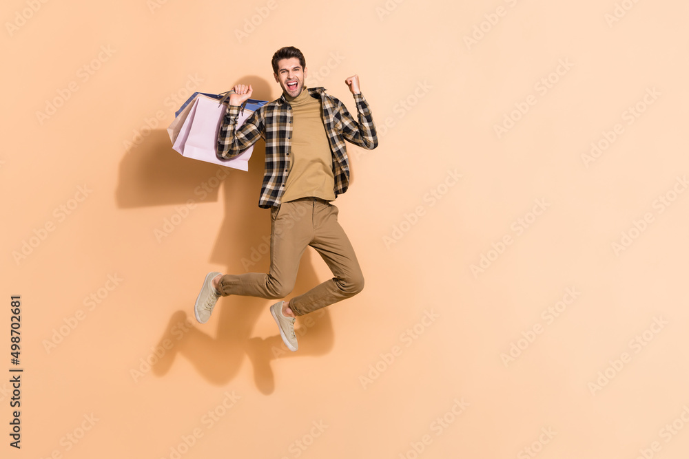 Full length body size view of attractive cheery lucky guy jumping holding bags having fun isolated on beige pastel color background Full length body size view of attractive cheery lucky guy jumping holding bags having fun isolated on beige pastel color background