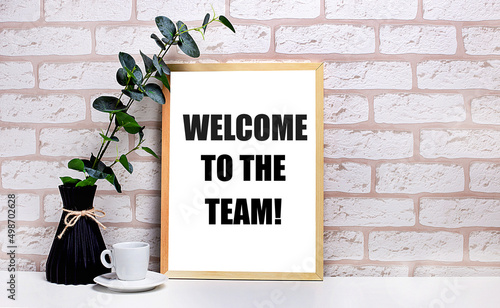 On a white table against a light brick wall, a branch of eucalyptus in a dark vase, a white cup and a light wooden frame with the text WELCOME TO THE TEAM. Home office interior.
