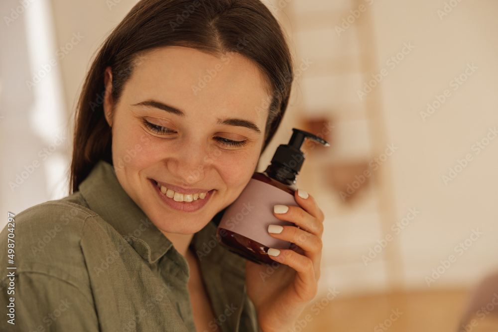 Close-up of young caucasian brunette girl smiling modestly while looking down indoors. Model in shirt holding body lotion. Skin care and hydration concept Close-up of young caucasian brunette girl smiling modestly while looking down indoors. Model in shirt holding body lotion. Skin care and hydration concept