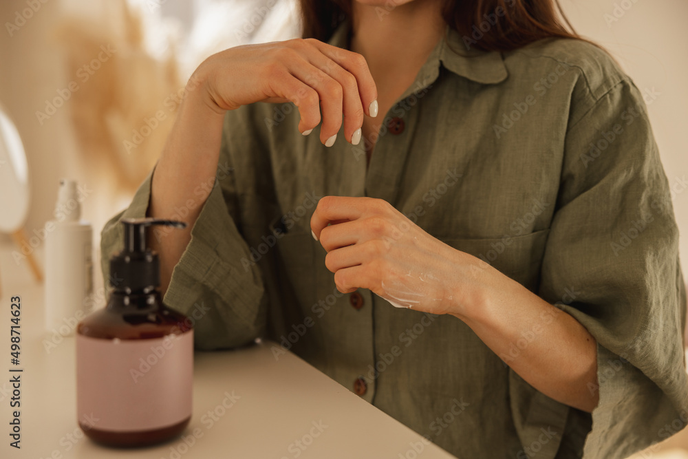Obraz premium Close-up of young caucasian woman's hands with clean skin applying cream at home. Girl in green shirt sits at table. Spa and beauty concept