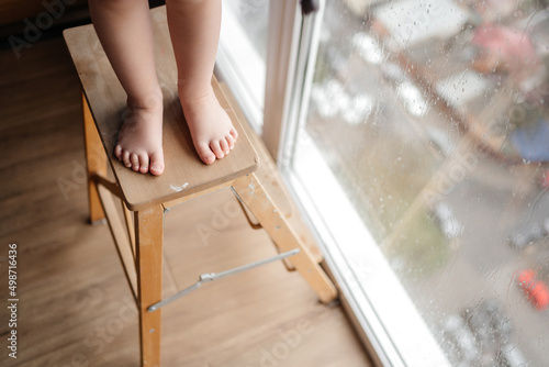 Legs of a small child on a ladder chair against a window with rainy weather. Attention Danger and children's interest in windows.