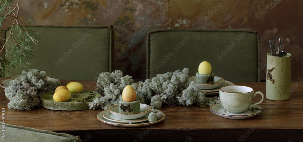 Table setting for Easter dinner decorated with forest moss, eggs coloured with matcha tea in moss green cement tray. Solid oak table against clay textured wall.