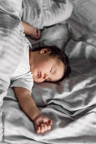 Portrait of little baby in sleepwear lying under gray blanket on bed at home. Sleeping infant child in bedroom, see sweet dream. Grey background, free copy space. Childcare and healthy sleep concept.