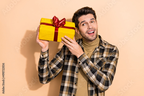 Portrait of attractive cheerful dreamy guy holding giftbox guessing what is inside isolated over beige pastel color background