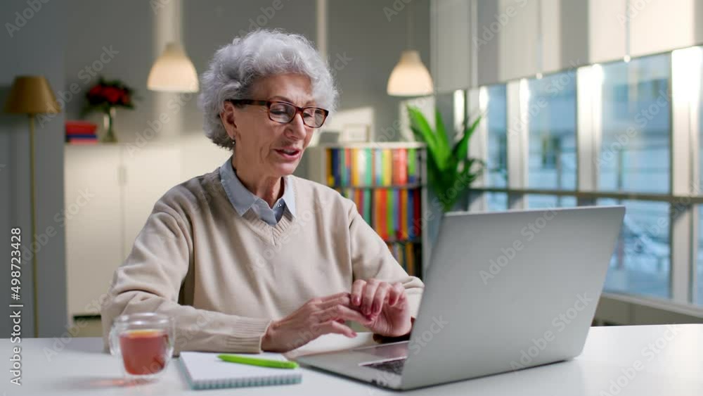 Senior woman having video call on laptop sitting at desk in modern living room Senior woman having video call on laptop sitting at desk in modern living room