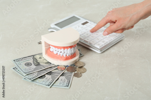 Woman using calculator near educational dental typodont model and money at light grey table, closeup. Expensive treatment photo