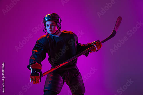 Portrait of boy, child, hockey player in protective uniform isolated over purple studio background in neon light © master1305