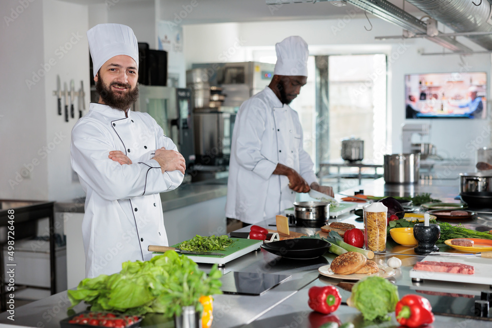 Smiling confident head chef standing in restaurant professional kitchen while looking at camera after recipe preparation. Skilled cook with arms crossed preparing dinner service ingredients. Smiling confident head chef standing in restaurant professional kitchen while looking at camera after recipe preparation. Skilled cook with arms crossed preparing dinner service ingredients.