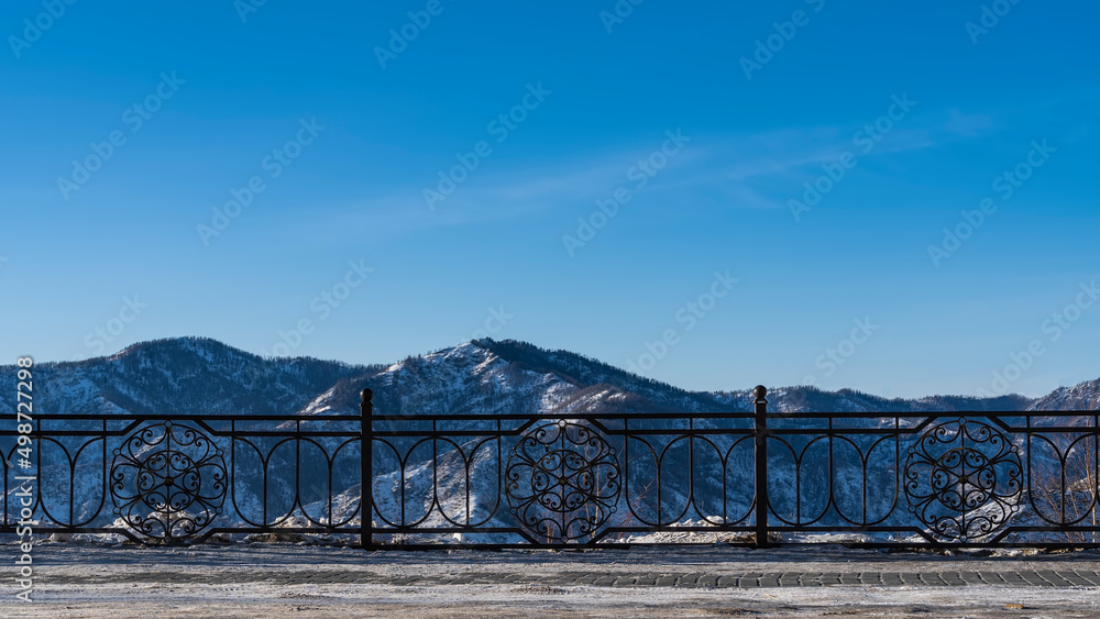 A metal fence is installed along the road. Through the openwork bars, snow-capped mountains can be seen against the blue sky. Copy space. Altai A metal fence is installed along the road. Through the openwork bars, snow-capped mountains can be seen against the blue sky. Copy space. Altai