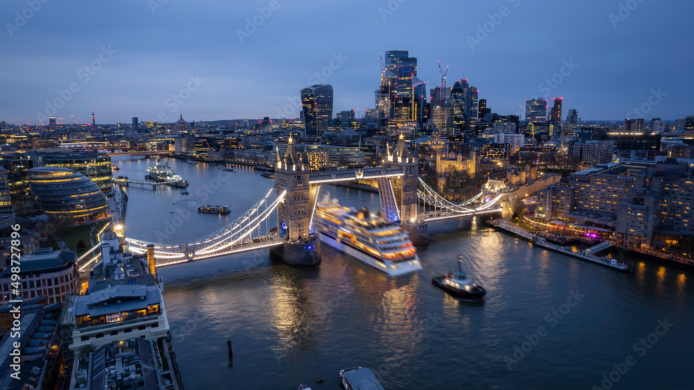 Aerial view of the skyline of London with a motion blurred cruise ship passing under the lifted Tower Bridge during dusk, England
