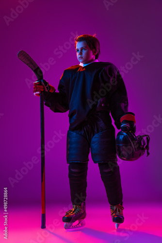 Full-length portrait of little boy, child, hockey player in special uniform posing with stick isolated over purple background in neon light © master1305