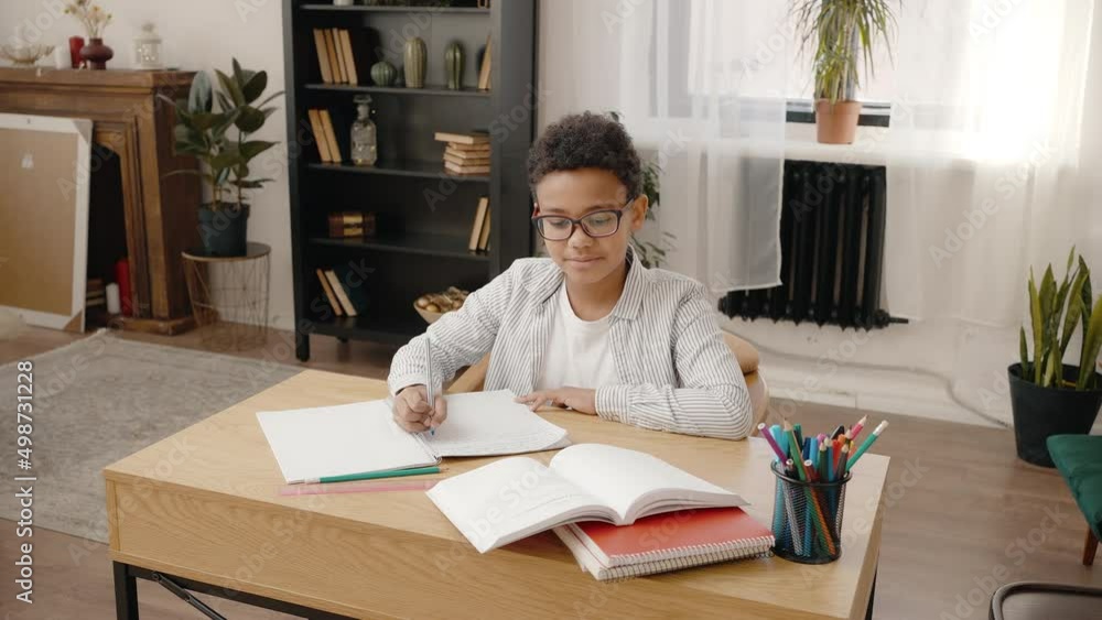 Domestic education. Happy african american boy in eyeglasses doing homework, writing in copy book, tracking shot Domestic education. Happy african american boy in eyeglasses doing homework, writing in copy book, tracking shot