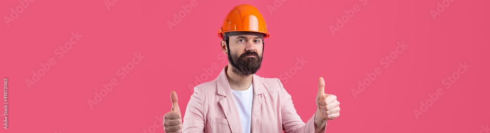 Portrait of young construction engineer wear orange hard hat, in a pink jacket standing on red studio background. The man points with his hand. Portrait of young construction engineer wear orange hard hat, in a pink jacket standing on red studio background. The man points with his hand.