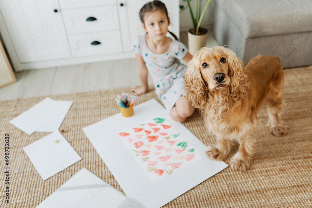 A girl draws hearts for his mother sitting on carpet floor in living room, cocker spaniel dog sitting nearby