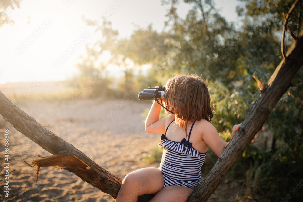 Cute european child girl in a bathing suit with binoculars on a tree. Summer holidays and adventures. Jungle games on the sea coast.