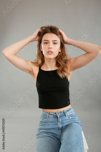 Beautiful girl model appearance in light jeans and black T-shirt on a gray studio background. Girl model sitting on a white chair. Beautiful blonde shows poses for a photo shoot  © Anhelina Tyshkovets