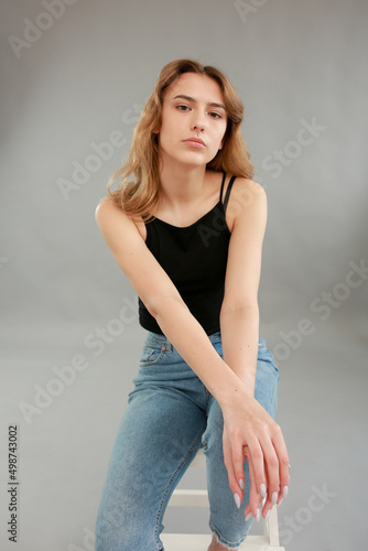 Beautiful girl model appearance in light jeans and black T-shirt on a gray studio background. Girl model sitting on a white chair. Beautiful blonde shows poses for a photo shoot 