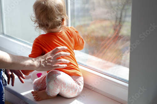 baby girl sitting on windowsill in front of the window glass at sunny day, mother holds child, natural dirty glass