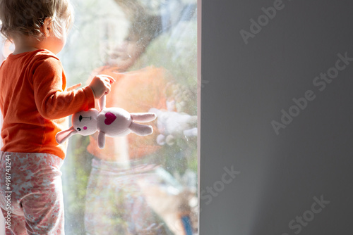 baby girl standing in front of the window glass at sunny day, holding rabbit toy with heart, natural dirty glass