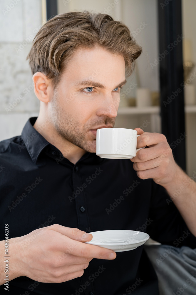 man in black shirt holding saucer and drinking coffee from white cup. man in black shirt holding saucer and drinking coffee from white cup.