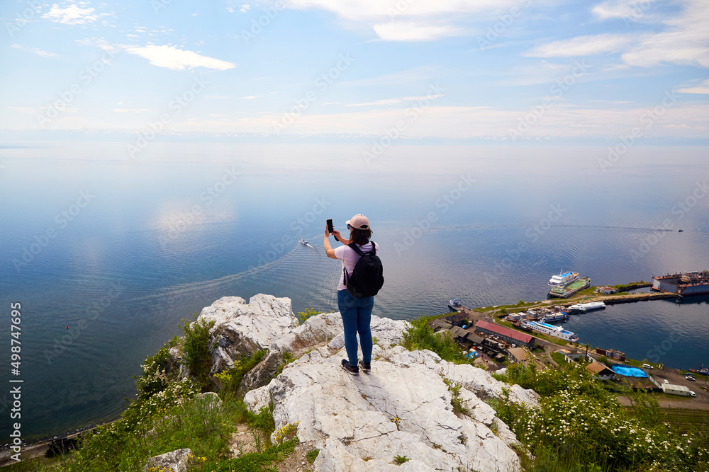 Fototapeta premium A female traveler stands on a rock near the port of Baikal and photographs Lake Baikal on a summer day. The concept of freedom, hiking.