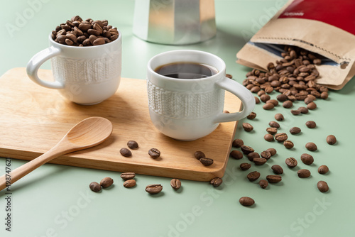 Cup of freshly brewed coffee, spoon and scattered coffee beans on a pastel green background. Whole organic coffee beans spilled out of a brown paper bag. Making tasty strong coffee concept.