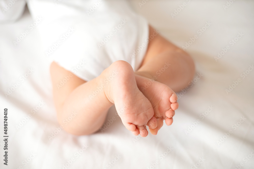 The legs of the baby in a white bodysuit on the white background of the bed. Light colors. Lifestyle. The legs of the baby in a white bodysuit on the white background of the bed. Light colors. Lifestyle.