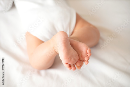The legs of the baby in a white bodysuit on the white background of the bed. Light colors. Lifestyle.