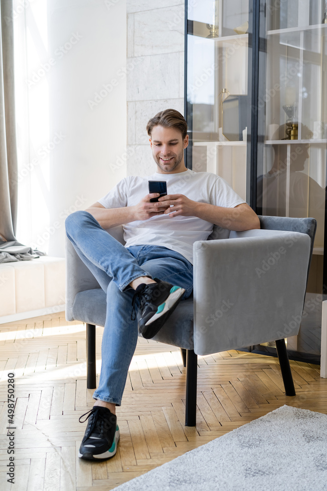 smiling man in jeans sitting in armchair with crossed legs and chatting on smartphone.