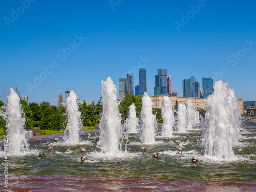 Jets of fountains on a sunny day against the backdrop of modern skyscrapers and a cloudless blue sky. Recreation area in Victory Park on Poklonnaya Hill in summer.