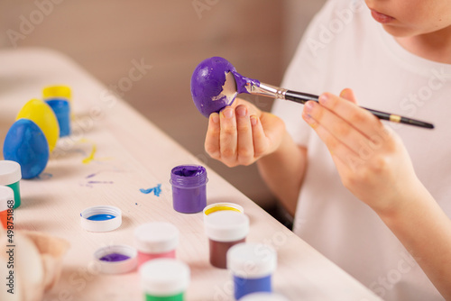 A Caucasian boy in a white t-shirt paints eggs to celebrate the Easter holiday with bright colors. Childish art  handmade culture concept. Boy with a brush in his hand paints an egg.