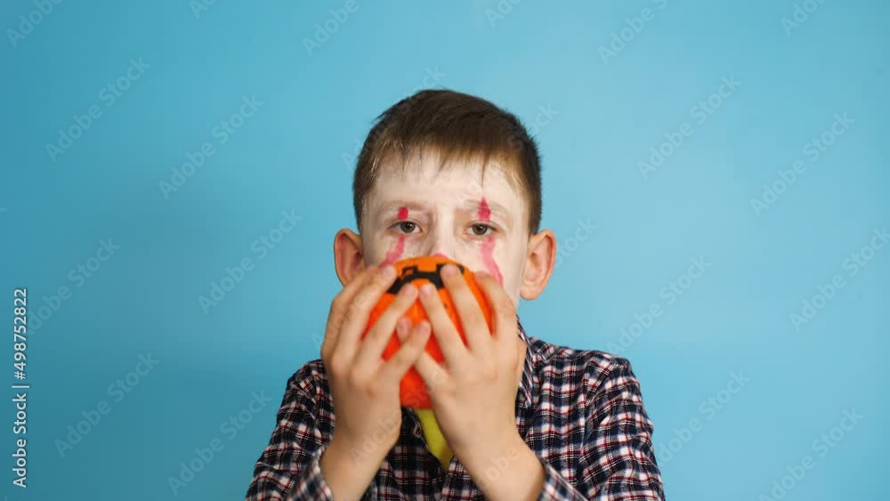 The child celebrates Halloween. Caucasian boy 7-8 years old in with a creepy make-up zombie holds a pumpkin lamp looks into the camera scares with a scary look. studio shooting blue background. The child celebrates Halloween. Caucasian boy 7-8 years old in with a creepy make-up zombie holds a pumpkin lamp looks into the camera scares with a scary look. studio shooting blue background.