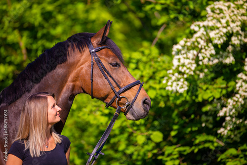 Portrait of a horse with a girl, the girl stands at neck level next to the horse and looks at him, in the background a flowering bush..