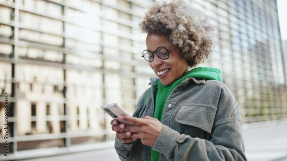 Handsome adult African woman wearing casual clothes typing by phone and walking outdoors Handsome adult African woman wearing casual clothes typing by phone and walking outdoors