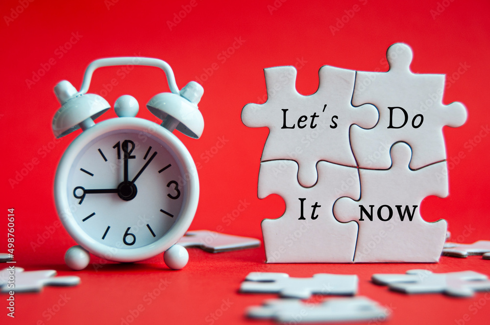 Let's do it now text on jigsaw puzzle with alarm clock pointing at 9 am on red cover background.