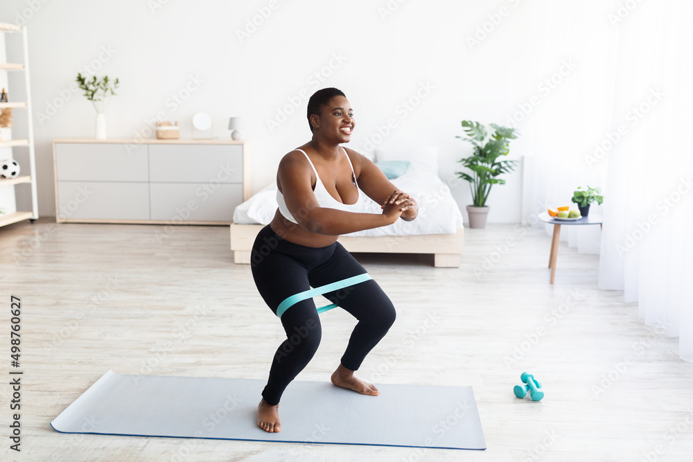 Chubby millennial black woman squatting with elastic band during domestic workout, free space Chubby millennial black woman squatting with elastic band during domestic workout, free space