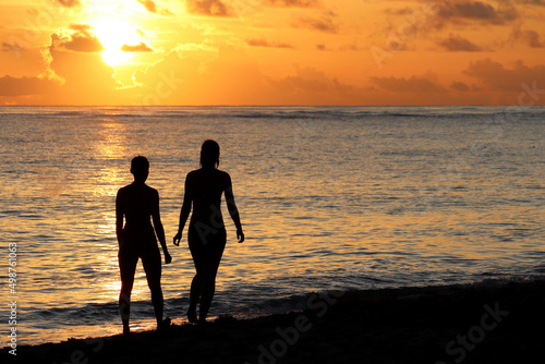 Silhouettes of two girls in bikini walking together by the sand on sea beach. Sunset on a coast, travel and holiday concept